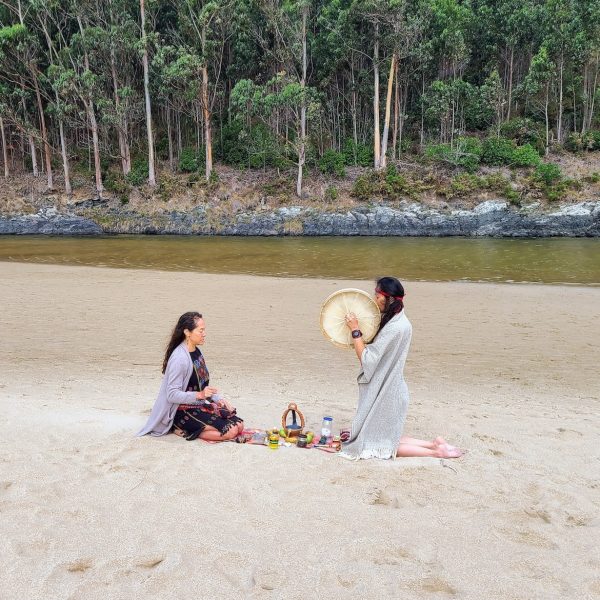 Two women engaged in a healing ritual on a sandy beach, with one playing a drum and the other sitting cross-legged surrounded by ritual items, against a backdrop of trees and a river.