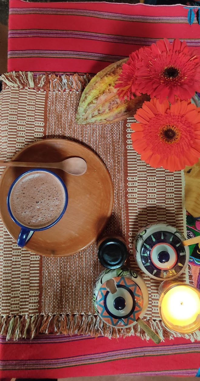 A cup of cacao on a woven mat, accompanied by a cacao pod, vibrant orange flowers, colorful bowls, and a lit candle, creating a cozy and inviting atmosphere.